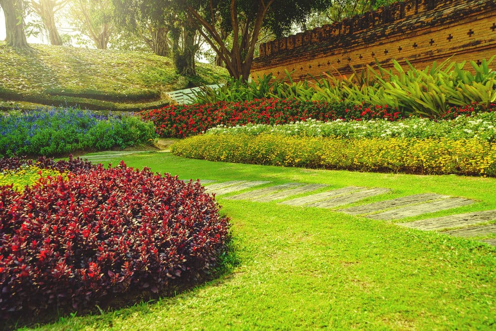 Vibrant garden scene with colorful flowers, lush greenery, and stone pathways under sunlight.